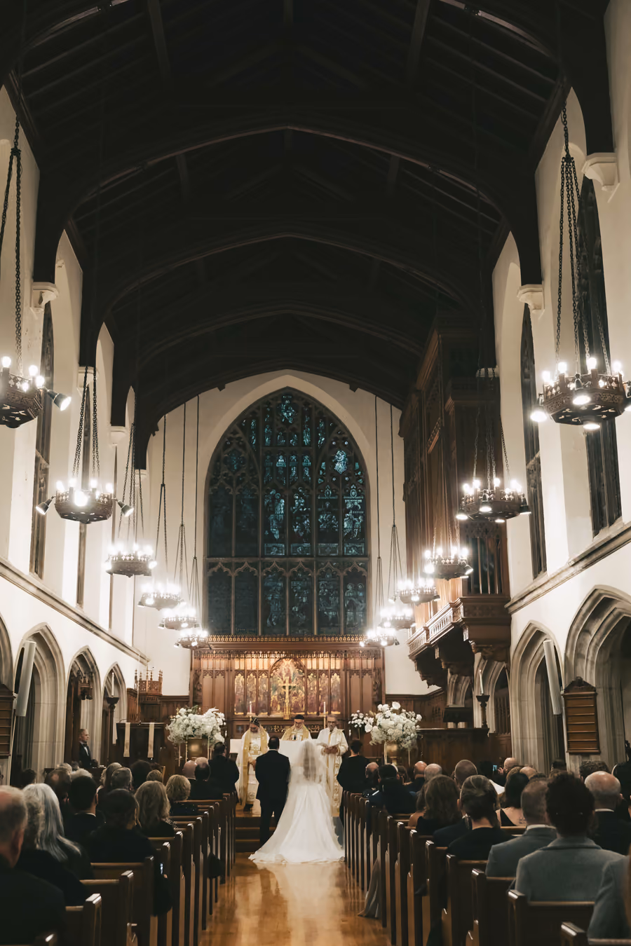 A church interior with gothic arches, hanging chandeliers, and stained glass above the aisle