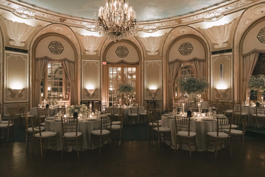 A grand ballroom reception with chandeliers, arched windows, and round dining tables