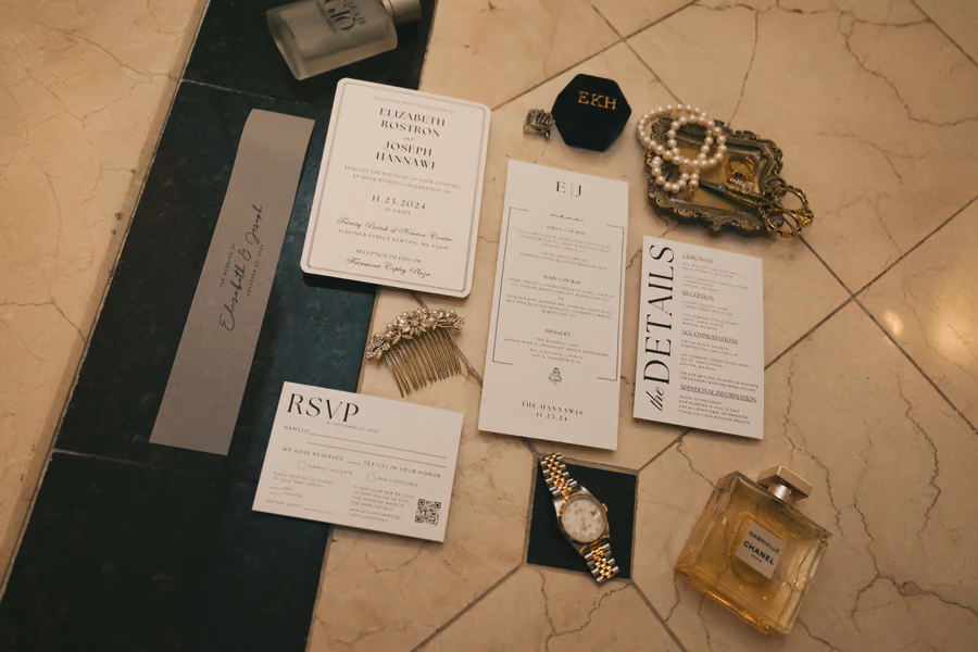 A wedding invitation suite laid out with rings, a watch, and perfume on a dark surface