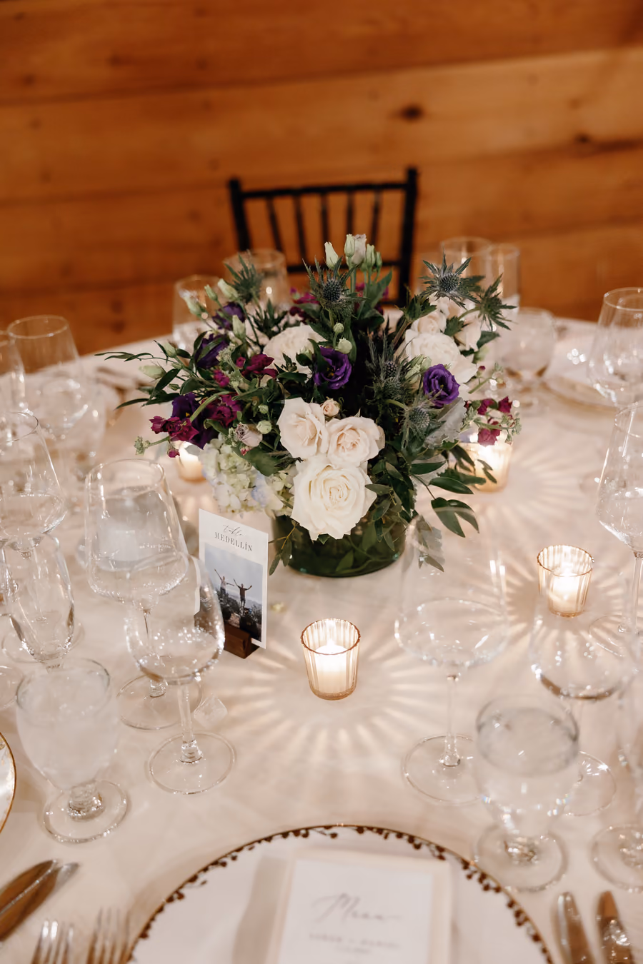 A reception centerpiece of white and purple flowers surrounded by glassware and votive candles