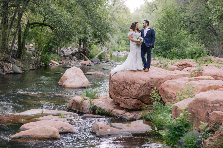 A couple embracing on red rocks beside a creek surrounded by green trees