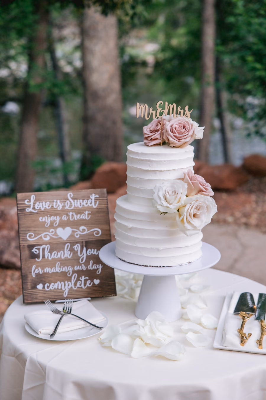 A tiered white wedding cake with blush roses beside a Love is Sweet dessert table sign
