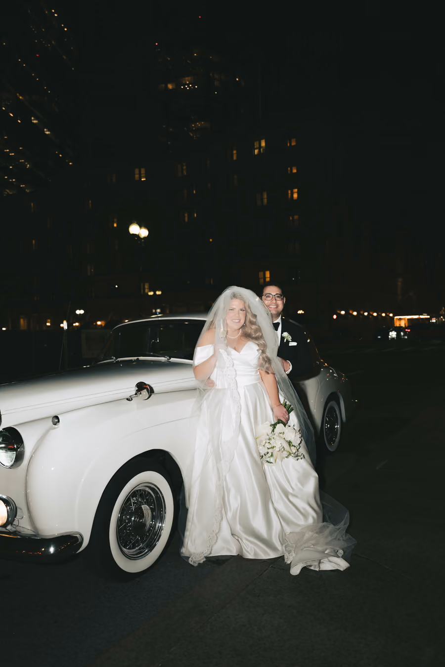 A couple standing beside a vintage white car at night with city lights behind them