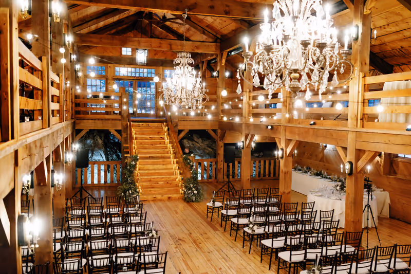 An empty wooden barn venue set up for a wedding ceremony with rows of dark chairs and chandeliers