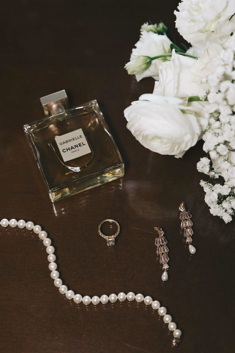 A flat lay of bridal details including a Chanel perfume bottle, white flowers, a pearl necklace, and earrings on a dark background