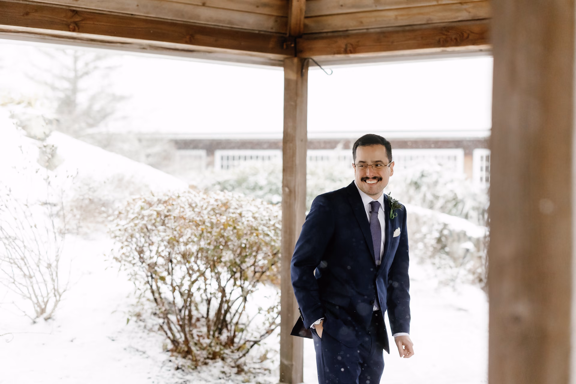 A groom in a blue suit smiling while standing outdoors in the snow under a wooden structure
