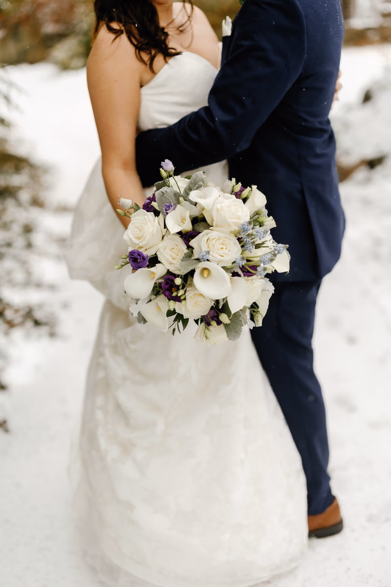 A close-up of a bride and groom embracing in the snow, with the bride holding a white and blue bouquet