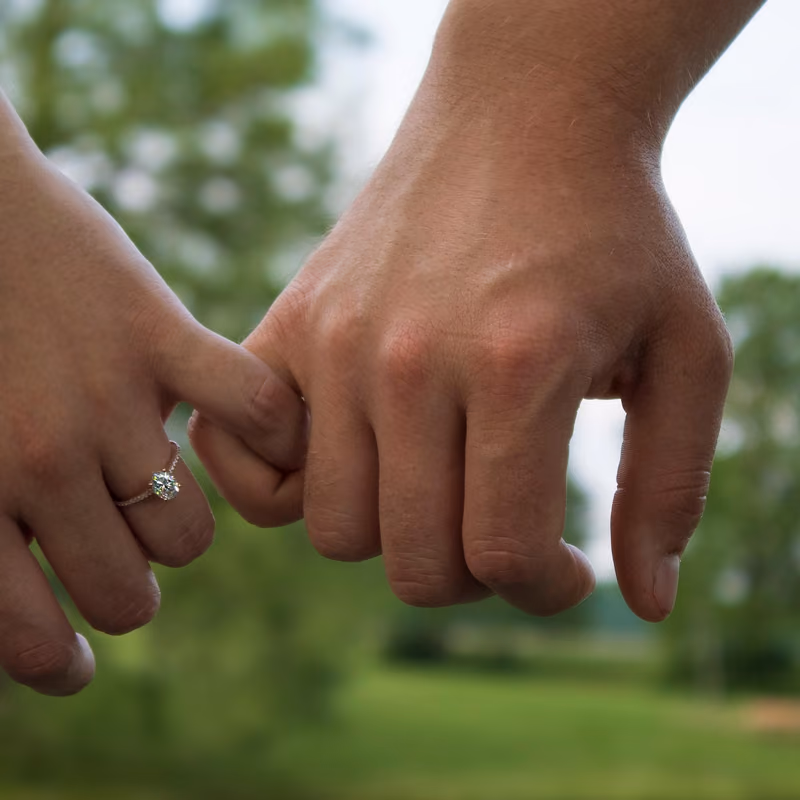 A close-up of a couple holding hands, showcasing an engagement ring against a grassy background