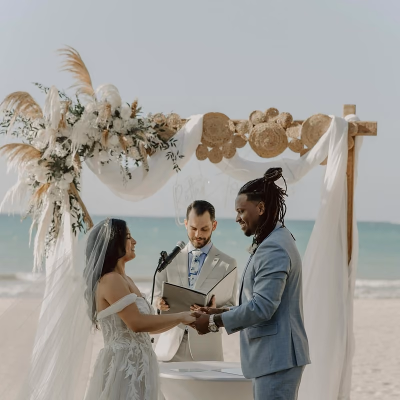 A couple exchanging vows under a floral arbor on a sandy beach