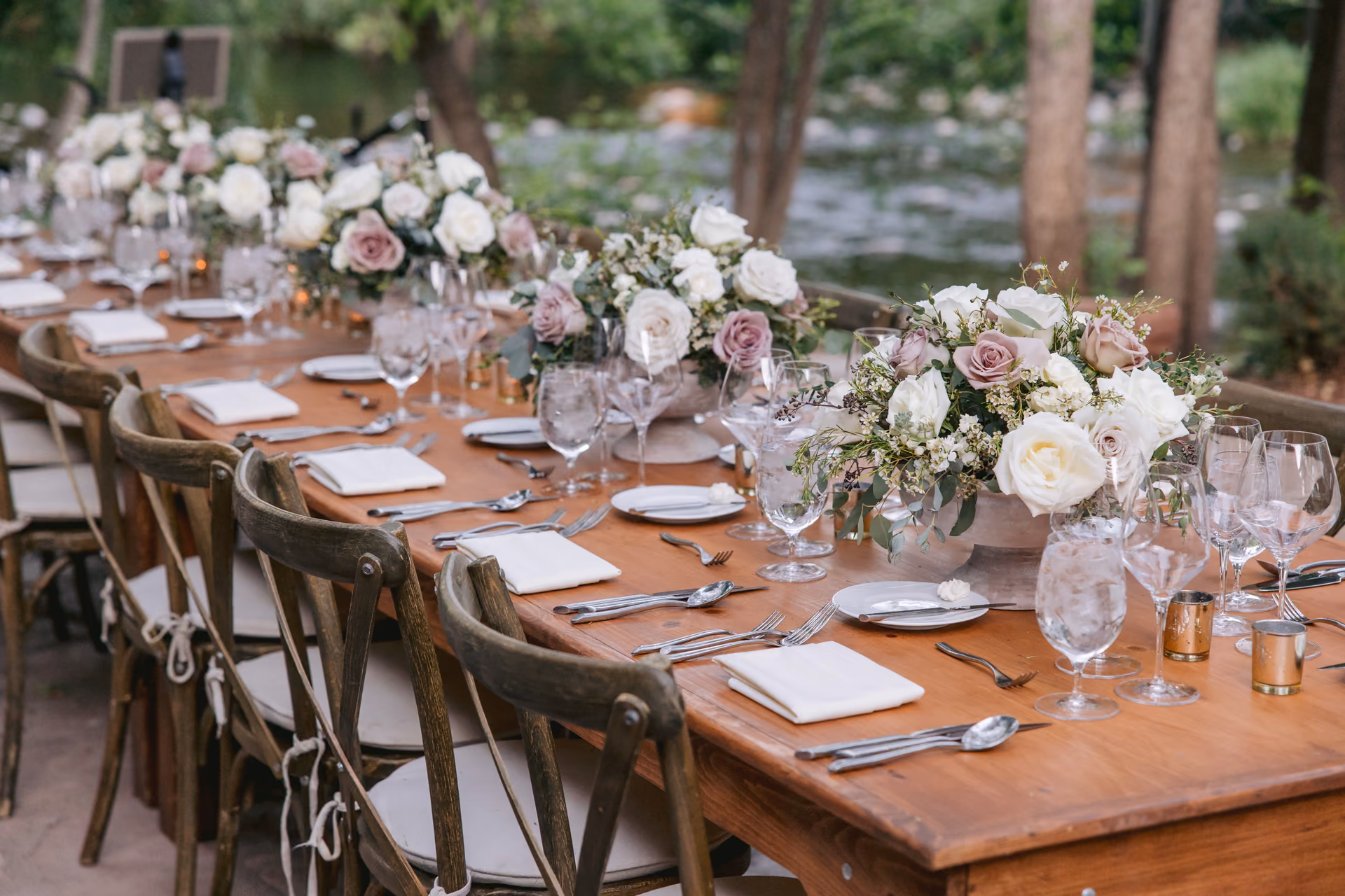 A long outdoor reception table set with blush florals and wooden chairs beside a creek
